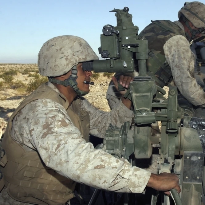 Cannoneer смотрит через прицелы M777 облегченного гаубица как другой crewman вставляет